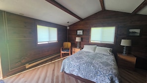 Un dormitorio con una cama, dos mesitas de noche, una silla y dos ventanas.