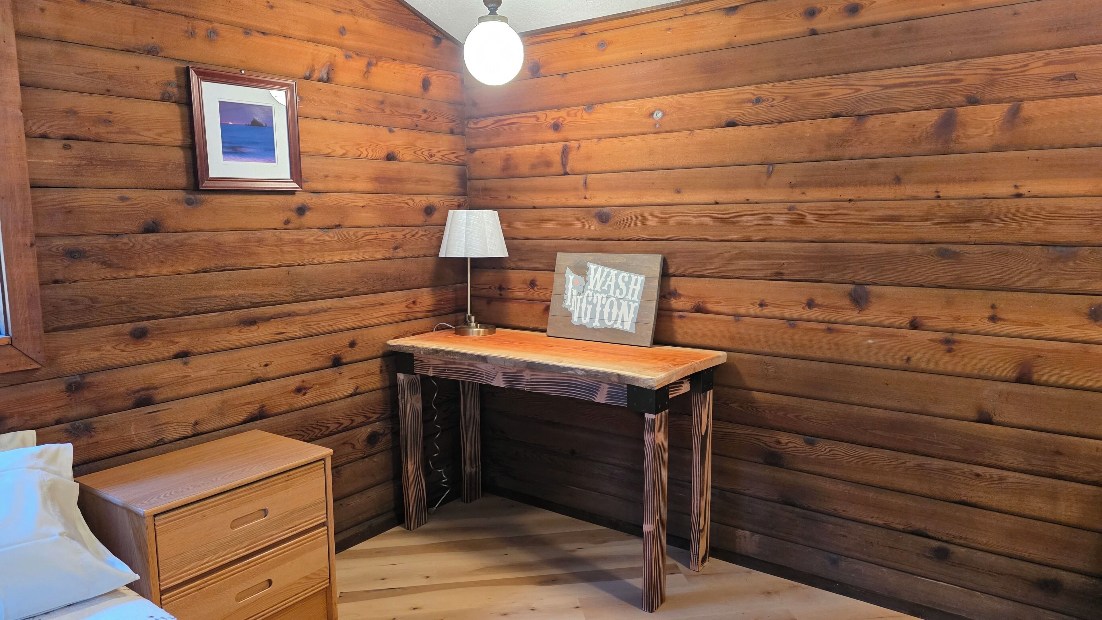 Una camera in una baita di legno con una scrivania, una lampada, un quadro incorniciato e un cartello con su scritto 'WASHington'.