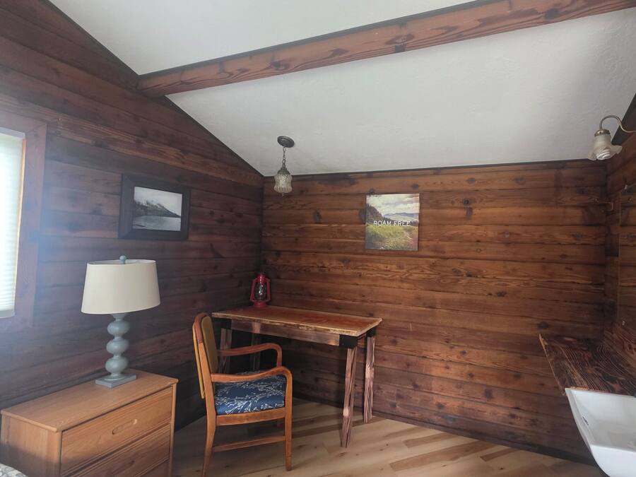Interior de una cabaña de madera con escritorio, silla, lámpara y un ventanal.