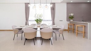 A modern dining area with a rectangular table, six chairs, pendant lighting, a window with curtains, and a kitchen area with stools. - Coco Villas - 3BR Deluxe Pool Villas  (Thep Krasattri)