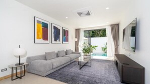 A modern living room with a grey sofa, a coffee table, a TV, and a large window with curtains. - Coco Villas - 3BR Deluxe Pool Villas  (Thep Krasattri)