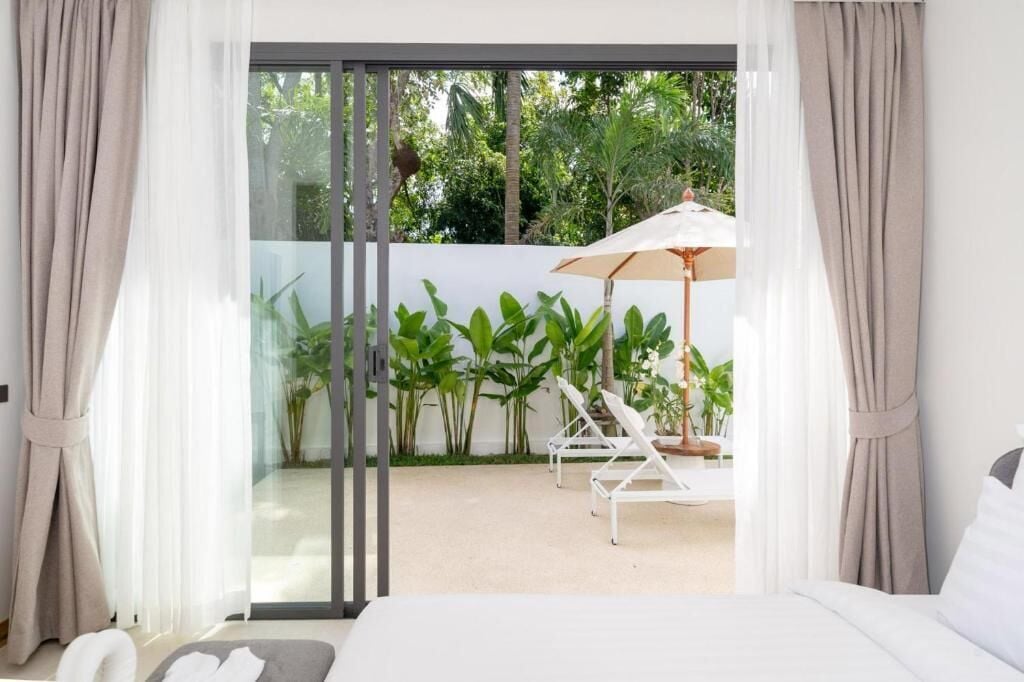 A bedroom with a large sliding glass door leading to an outdoor area with a white lounge chair and umbrella, surrounded by greenery.