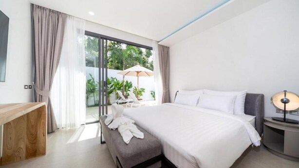 A modern hotel room with a large bed, a bedside table, a bench, and a view of the outdoors. - Coco Villas - 3BR Deluxe Pool Villas  (Thep Krasattri)