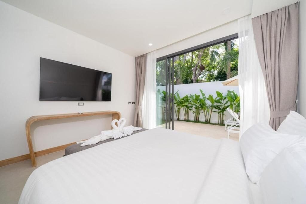 Quarto de hotel moderno com uma cama grande, televisão de tela plana na parede e uma porta de vidro deslizante que dá para uma varanda com plantas.