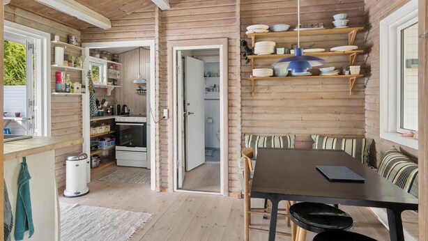 Interno di una baita in legno con zona pranzo, mensole con stoviglie e una cucina visibile attraverso una porta aperta.