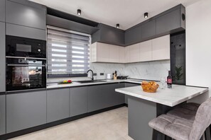 Eine moderne Küche mit grauen Schränken, integriertem Backofen, einem Fenster mit Jalousien, einer Marmorarbeitsplatte und einer Barbereich mit einem Stuhl.