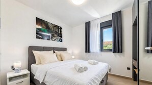 Een moderne hotelkamer met een bed, nachtkastje, schilderij, raam met gordijnen en uitzicht op de omgeving.