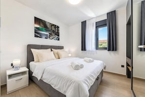 Ein modernes Hotelzimmer mit Bett, Nachttisch, einem Gemälde, einem Fenster mit Vorhängen und Blick ins Freie.