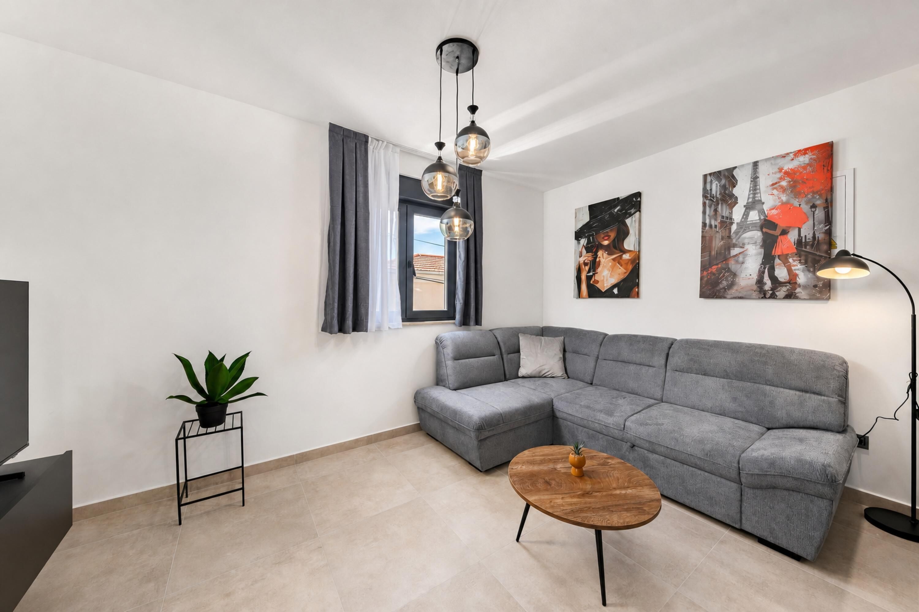 Una sala de estar moderna con un sofá seccional gris, una pequeña mesa de centro redonda de madera, una planta en maceta y obras de arte en las paredes.