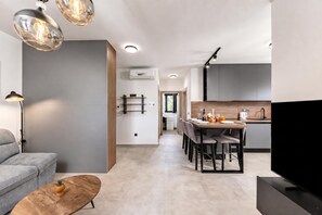 Ein modernes Wohnzimmer mit einer Couch, einem Couchtisch und einem Fernseher. Im Hintergrund ist ein Essbereich mit Tisch und Stühlen zu sehen.
