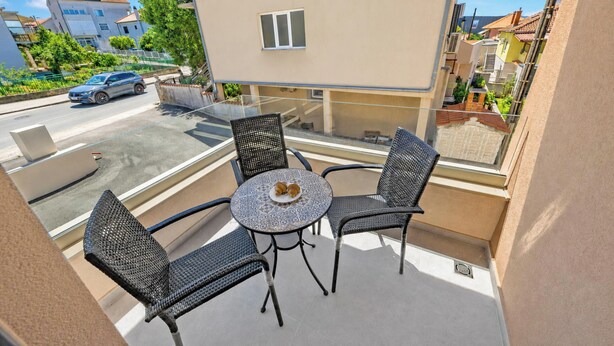 Een balkon met een glazen tafel en vier stoelen, met uitzicht op een straat met geparkeerde auto's en gebouwen.