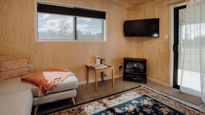 Ein gemütliches Zimmer mit einer Couch, einem kleinen Tisch mit einer Lampe, einem an der Wand befestigten Fernseher und einem großen Fenster mit Blick auf Bäume.