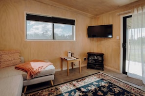 Ein gemütliches Zimmer mit einer Couch, einem kleinen Tisch mit einer Lampe, einem an der Wand befestigten Fernseher und einem großen Fenster mit Blick auf Bäume.
