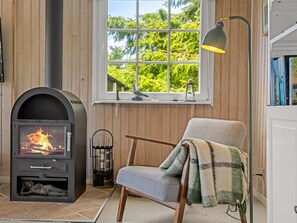 Ein gemütliches Zimmer mit Kamin, einem Sessel mit Decke, einem Fenster mit Blick auf Grünflächen und einem Bücherregal.