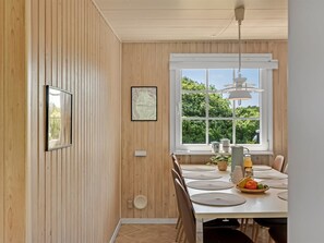 Un comedor con pared de madera, una ventana con vistas a la vegetación, una lámpara colgante y una mesa preparada con individuales y un frutero.