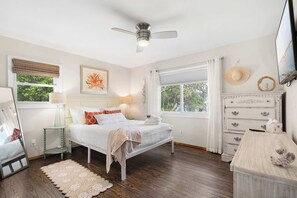 Ein Schlafzimmer mit Bett, Nachttischen, Deckenventilator, Fenster mit Vorhängen, Kommode und einem gerahmten Kunstwerk an der Wand.
