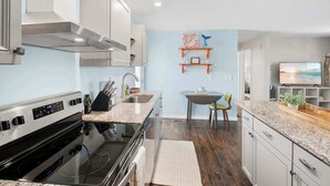 A modern kitchen with a built-in stove, stainless steel range hood, and a dining area with a table and chairs. - Walk to Surfside Beach Access, Golf Cart Add On (St. Augustine)