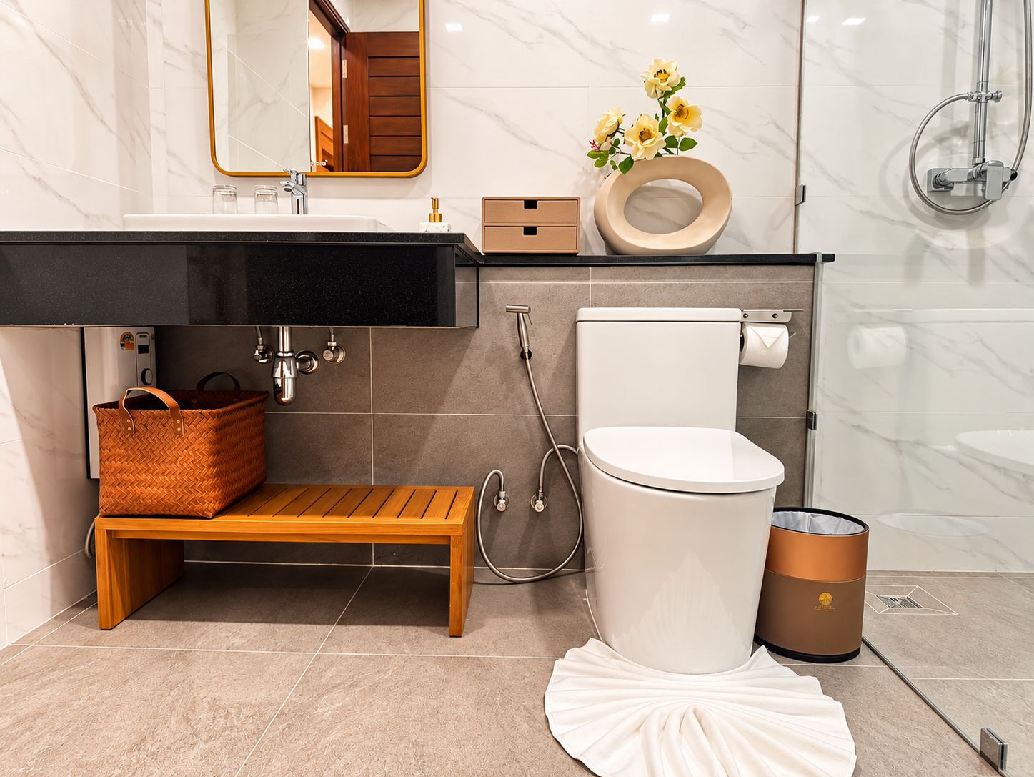 Un baño moderno con lavamanos de mármol, banco de madera, inodoro y un cubo de basura.