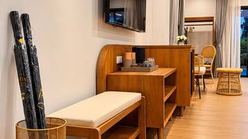 A modern hotel room with a wooden bench, a small desk, a television mounted on the wall, and a wicker chair.