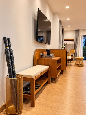 Une chambre d’hôtel moderne avec un banc en bois, un petit bureau, un téléviseur fixé au mur et un fauteuil en osier.