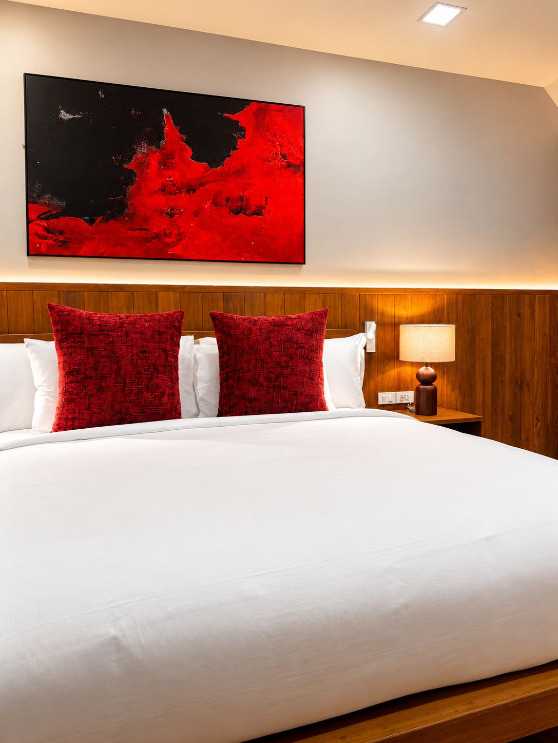Une chambre d’hôtel avec un grand lit, deux oreillers rouges, une couette blanche, une tête de lit en bois, une table de chevet avec une lampe et un tableau au mur.