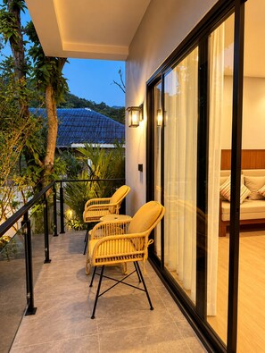 Un balcon avec des fauteuils en osier, une porte vitrée donnant sur une pièce avec un lit, et une vue sur une maison avec un toit bleu.