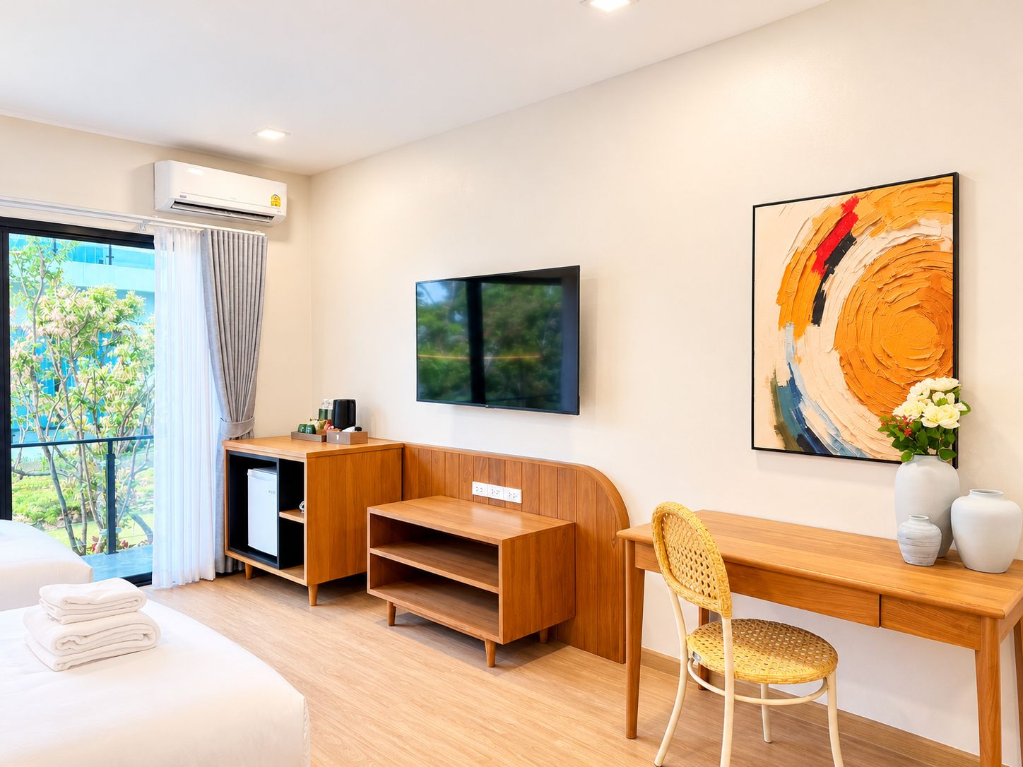 A modern hotel room with a wooden desk, a television, a large abstract painting, and a view of greenery outside.