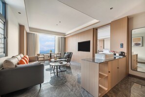 A modern hotel room with a sofa, a dining table, a TV, and a kitchen area with a sink.