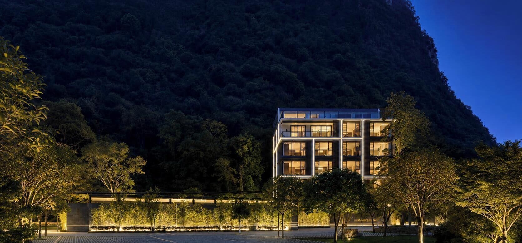 A modern building with multiple floors, surrounded by trees and a mountain backdrop.