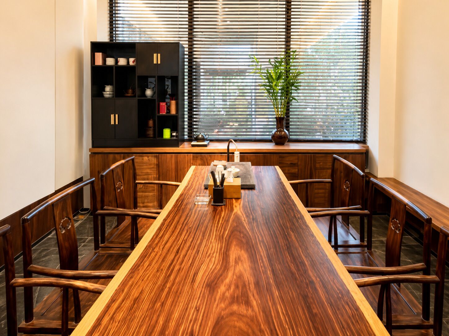 Un comedor con una mesa de madera larga, sillas, una estantería negra y una ventana con persianas.