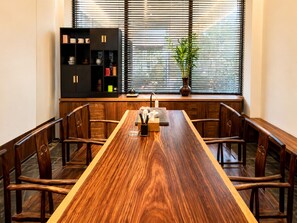 Un comedor con una mesa de madera larga, sillas, una estantería negra y una ventana con persianas.
