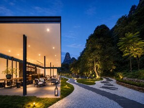 A modern building with a glass facade, outdoor seating area, and a pathway leading to a mountain view. - The Floral Dreamscape Retreat -Yulong Romance Park Branch-Tickets Reservation & Day Tour Arrangement (Guilin)