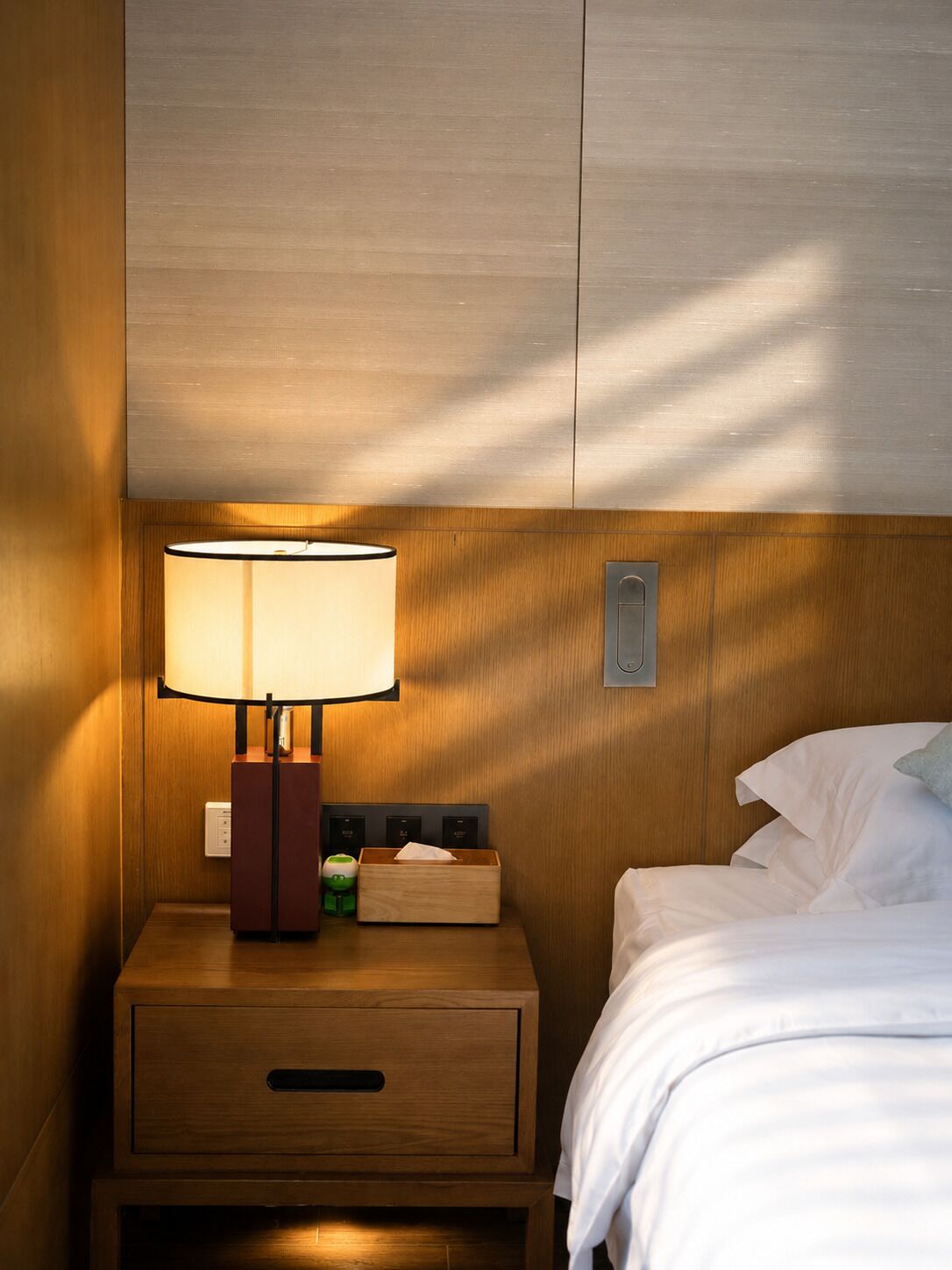 Een hotelkamer met een houten nachtkastje, een lamp, een zakdoosje, een stopcontact en een bed met witte lakens.
