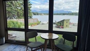 A room with a large window overlooking a lake, two chairs, a small round table, and a wooden deck with a shed. - 4BR/4BA Lakefront Lodge- mountain views, wildlife, canoeing- 9 min to airport (Anchorage)