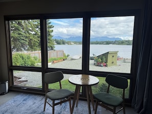 Ein Zimmer mit großem Fenster mit Blick auf einen See, zwei Stühle, ein kleiner runder Tisch und eine Holzterrasse mit einem Schuppen.