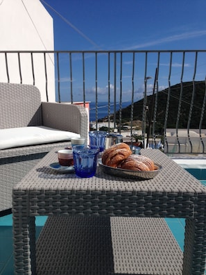 A wicker table with a tray of croissants and blue glasses, set against a backdrop of a clear sky and a mountainous landscape.