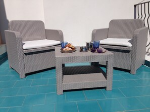 Two wicker chairs with white cushions, a small table with a tray of food and drinks, and a blue tiled floor.