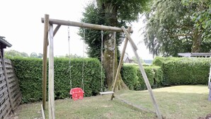 Een houten schommel in een gazon, met een rode plastic emmer en een haag op de achtergrond.