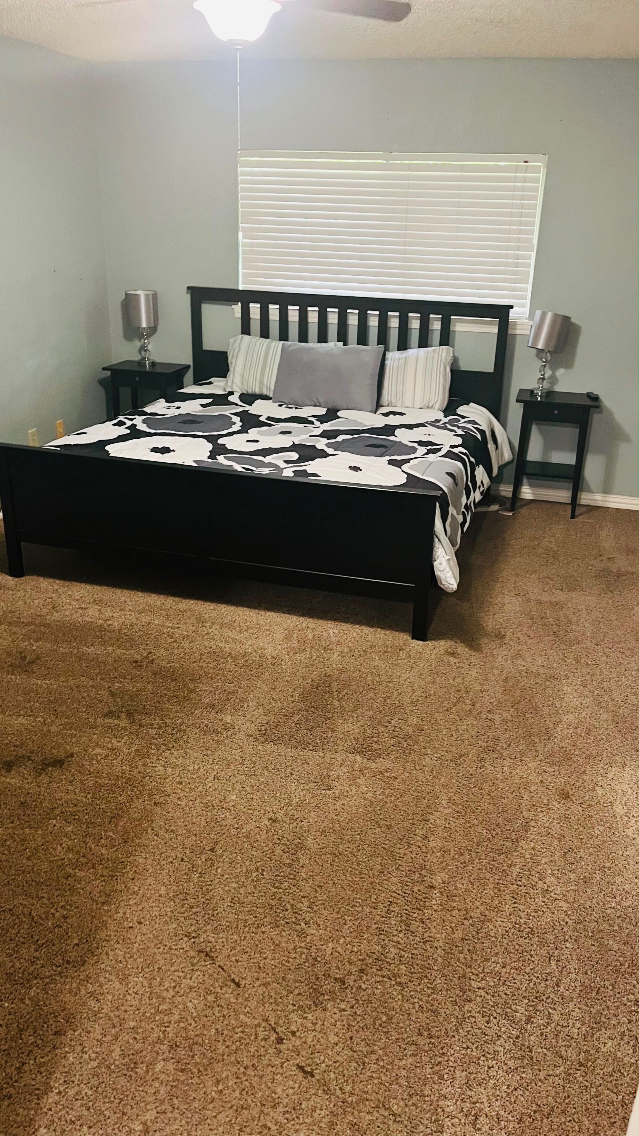A bedroom with a bed, two bedside tables, a window with blinds, and a carpeted floor.