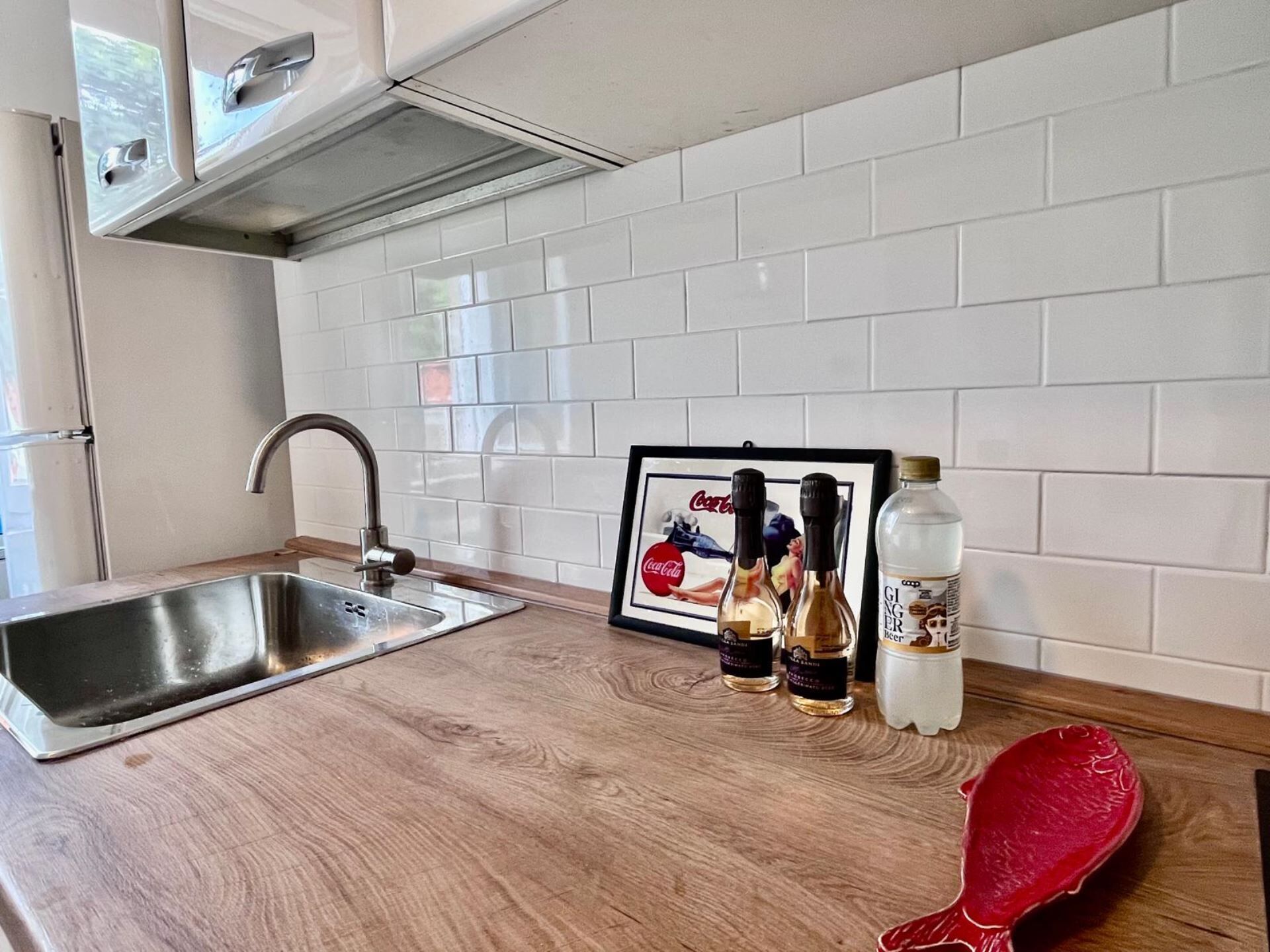 A kitchen with a stainless steel sink, a framed picture, and three bottles of ginger beer on the counter.