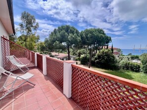 A balcony with a red lattice fence, a white lounge chair, and a view of the sea and trees. - Villa Blu di Norsi: 100 steps to sea and peace (Capoliveri)
