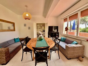 A cozy living room with a wooden dining table, black chairs, a woven sofa, a television, and a large window with a view. - Villa Blu di Norsi: 100 steps to sea and peace (Capoliveri)