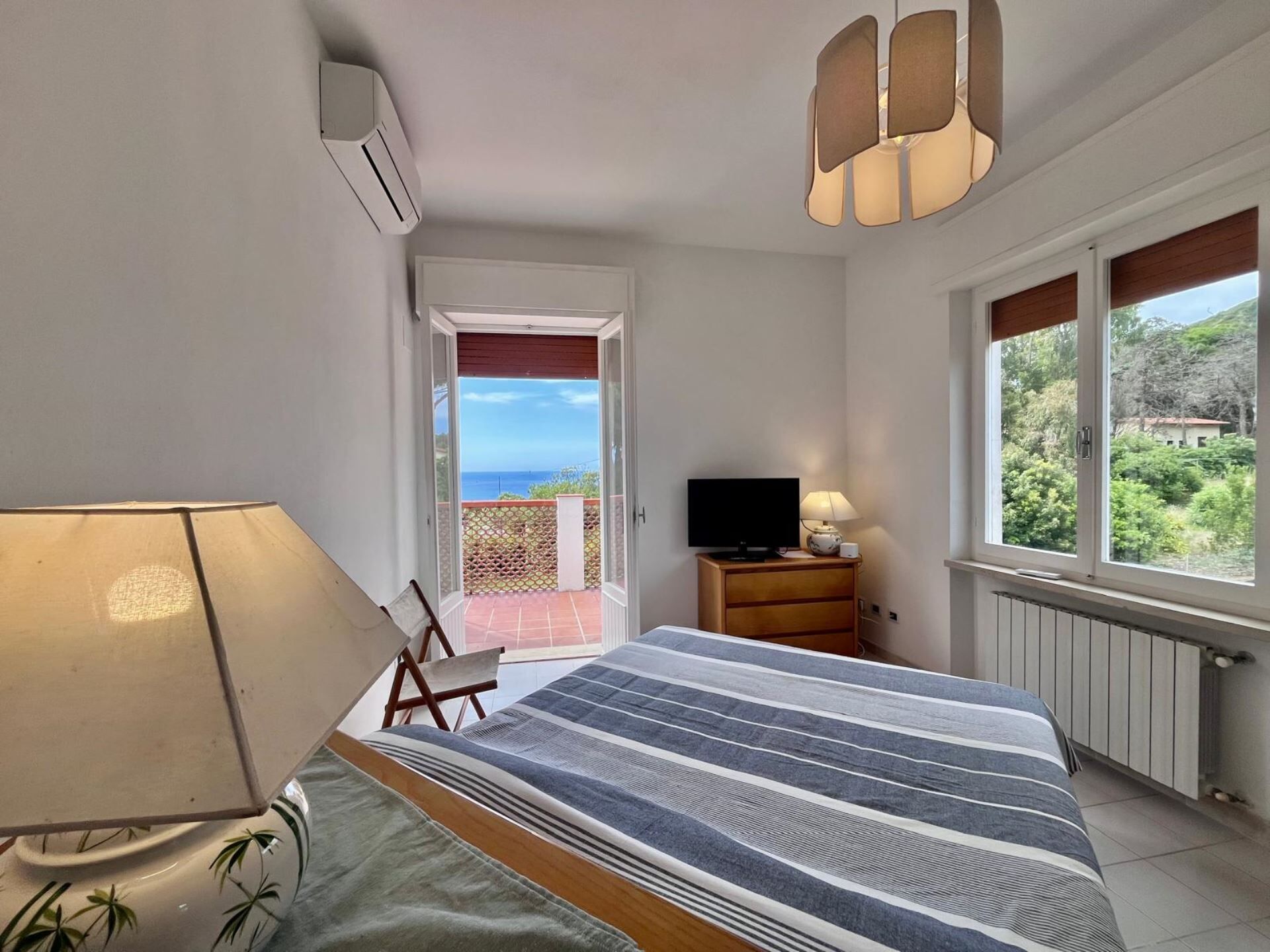 A bedroom with a bed, a TV, a lamp, a window with a view, and an air conditioning unit.