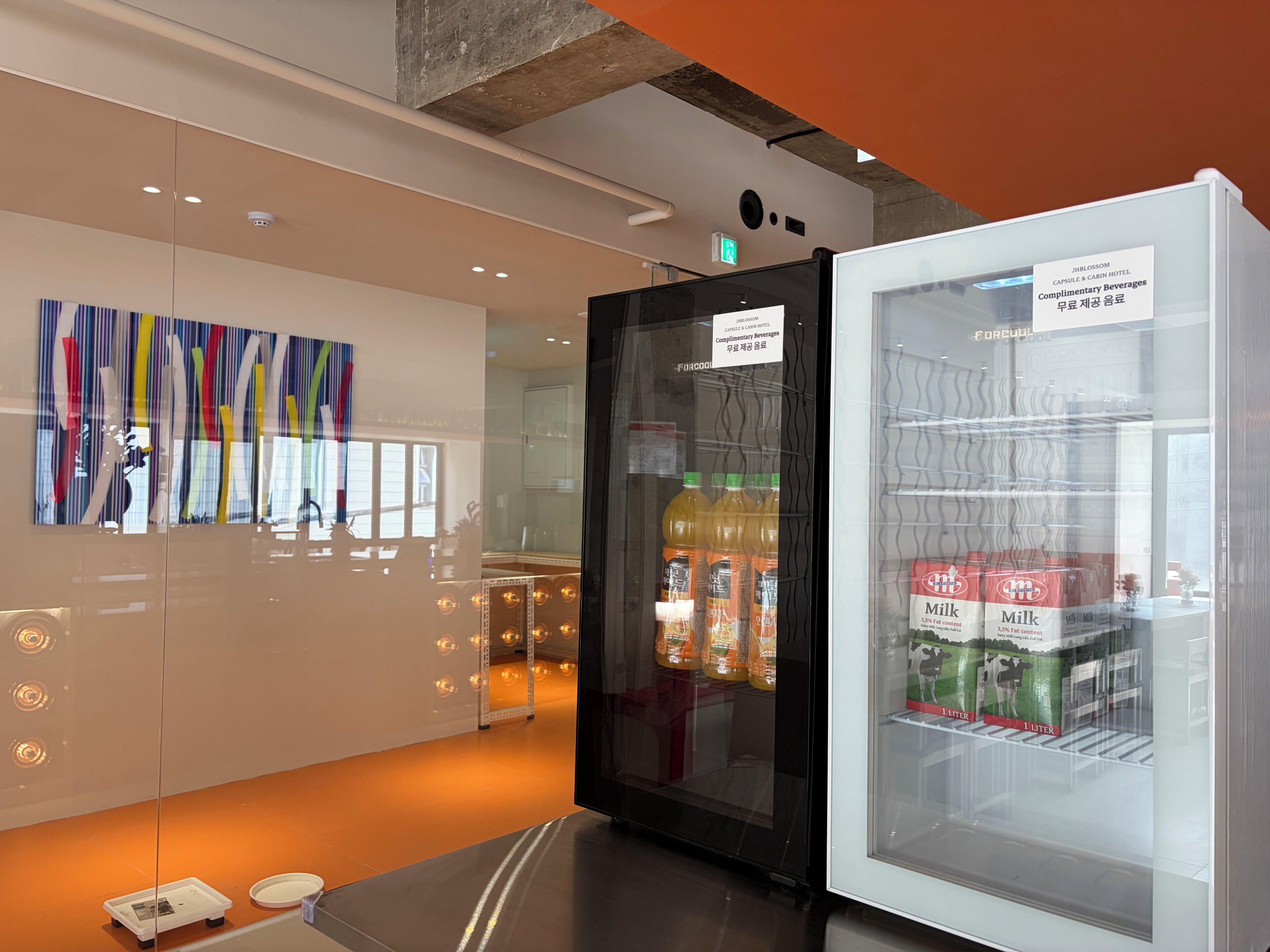 A hotel room with a refrigerator stocked with beverages, a glass display case with milk cartons, and a colorful abstract painting on the wall.