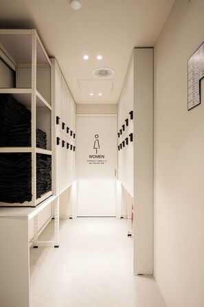 A corridor with storage shelves on both sides, a door with a women's restroom sign, and a ceiling with recessed lighting. - Jh blossom capsule & cabin hotel (Busan)