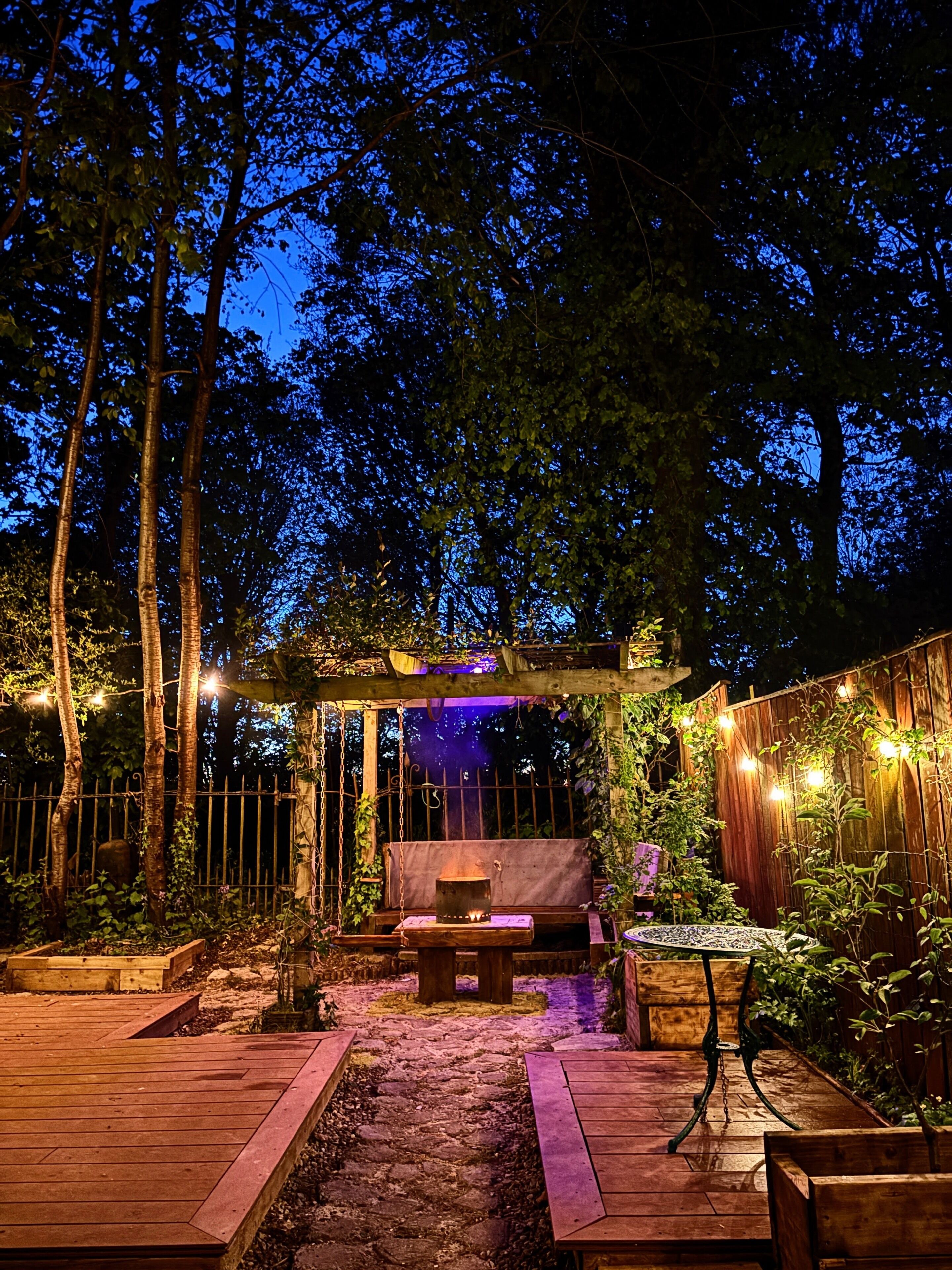 Un patio al aire libre con un deck de madera, una zona de estar con mesa y sillas, y una pérgola con luces colgantes.