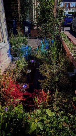 Un jardín con un estanque, diversas plantas y un contenedor de reciclaje.