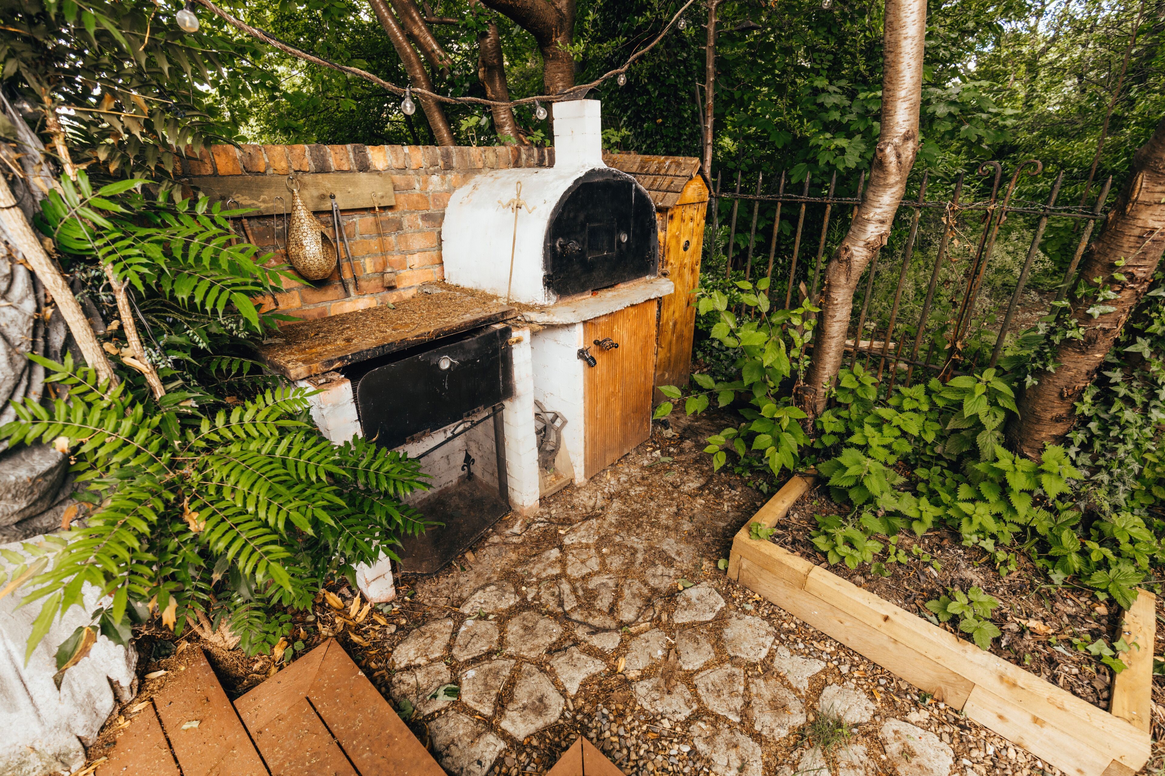 A brick oven with a wooden door, surrounded by greenery and a stone pathway.