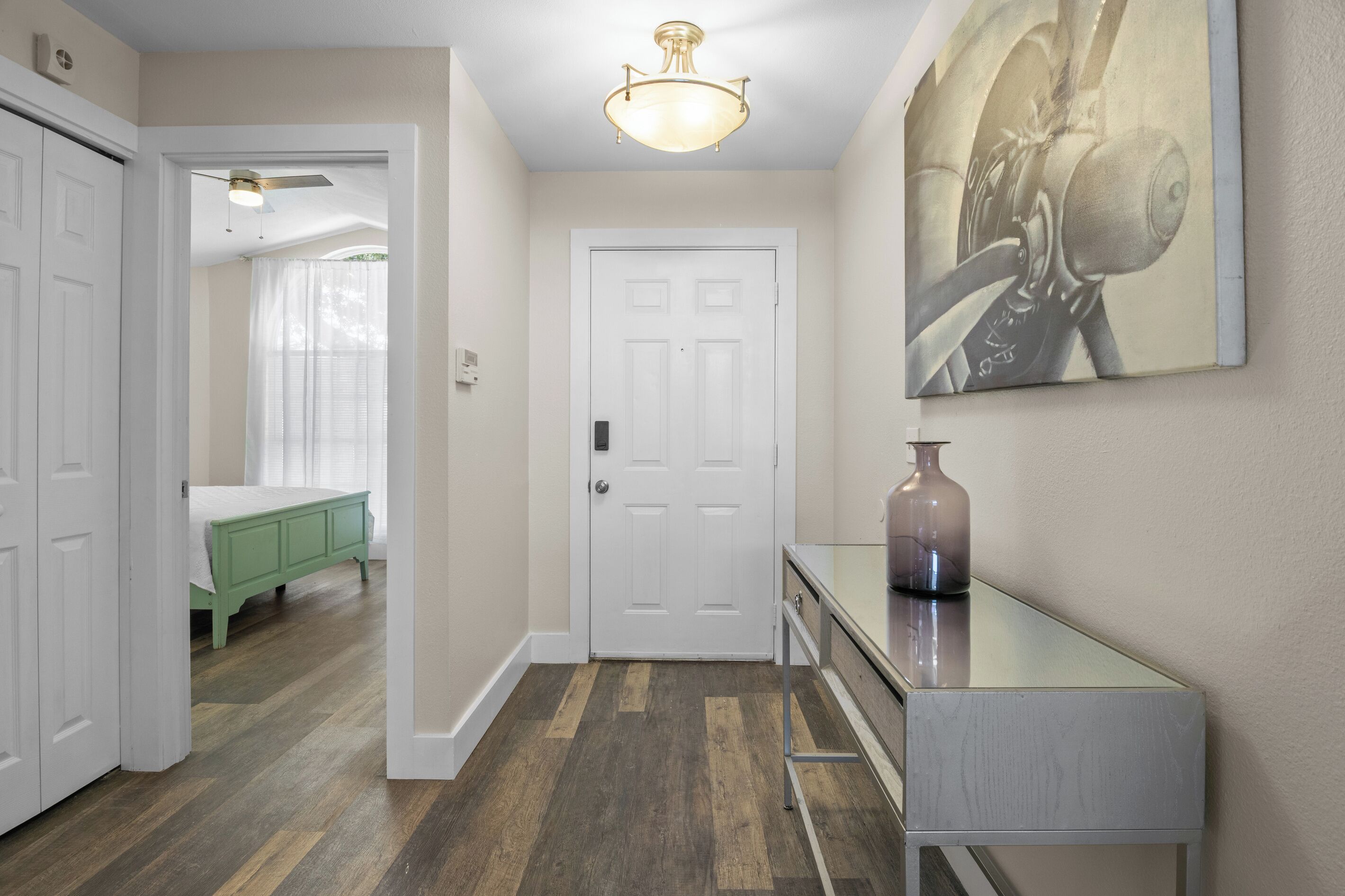 A hallway with a white door, a bed with green bedding, a glass table with a vase, and a ceiling light fixture.