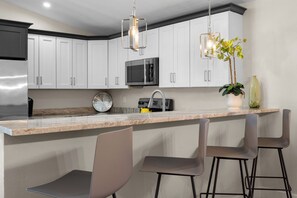 A modern kitchen with white cabinets, a stainless steel refrigerator, a microwave, a clock, and a bar area with chairs. - The Aurelia House | Elegant South Lakeland (Lakeland)